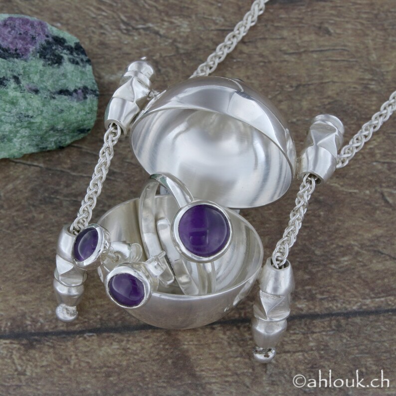 May include: A silver necklace with a round, hinged locket that opens to reveal three rings with purple gemstones. The locket is suspended from a silver chain.