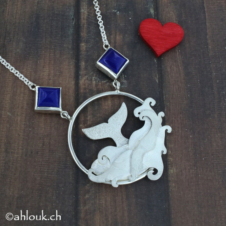 May include: A silver necklace with a whale tail and wave design pendant. The pendant is surrounded by a circle and features two blue gemstone accents.
