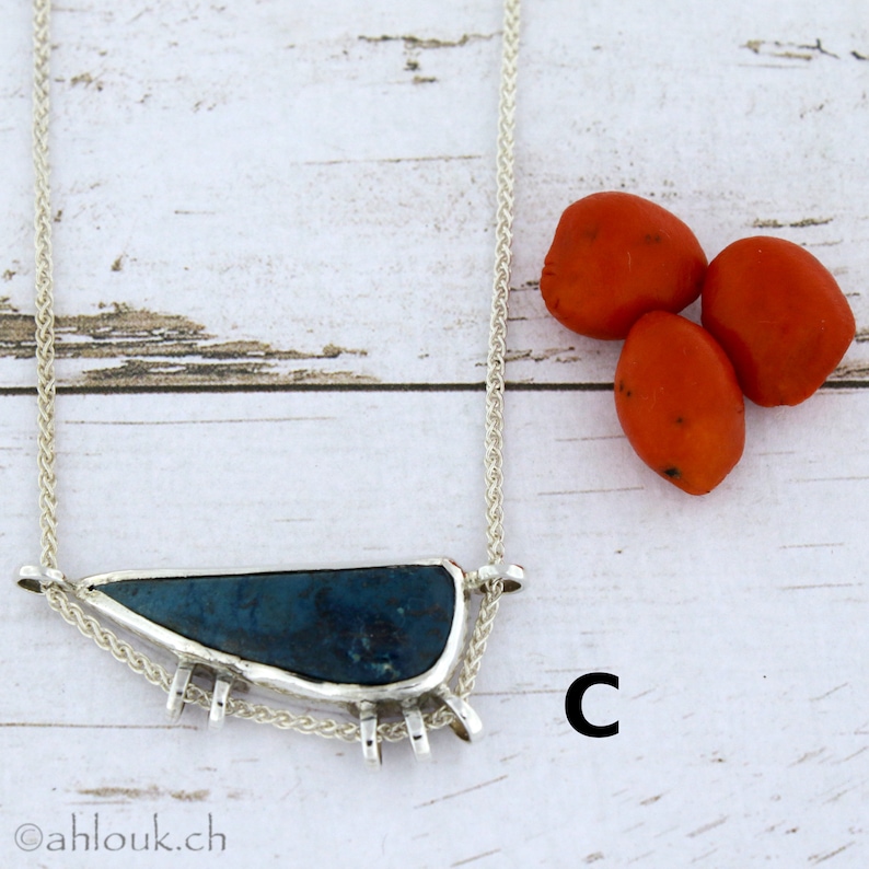 Puede incluir: Un collar de plata con un colgante de piedra azul y una cadena de cuerda. El colgante est&aacute; engastado en un marco de plata con peque&ntilde;as p&uacute;as alrededor del borde. El collar est&aacute; sobre una superficie de madera blanca con tres bayas rojas.