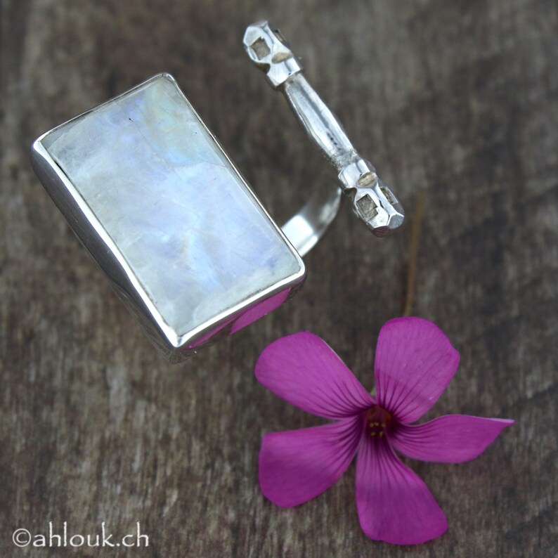 May include: A silver ring featuring a rectangular moonstone and a decorative silver band. The ring is displayed on a wooden surface with a pink flower.