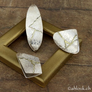 May include: Four silver-toned jewelry pieces with a textured finish and gold-colored accents. The pieces include an oval pendant, a triangular ring, and two square-shaped rings. The jewelry is displayed on a wooden surface within a gold-colored frame.