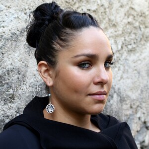 May include: A woman with dark hair styled in a bun wears a black top and silver earrings with a turquoise stone and a geometric design.