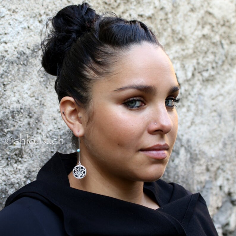 May include: A woman with dark hair styled in a bun wears a black top and silver earrings with a turquoise stone and a geometric design.