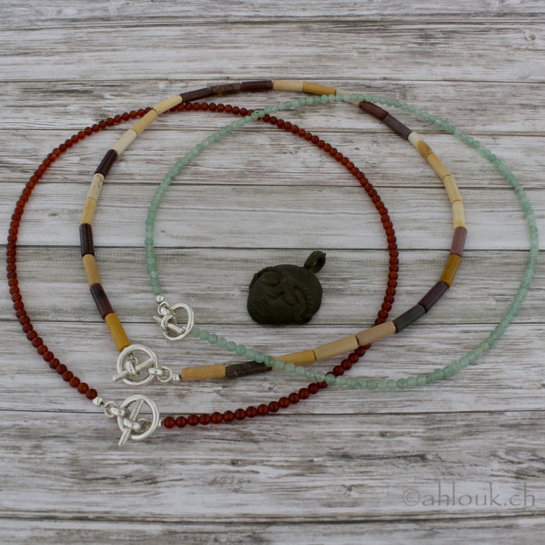 May include: Two necklaces with toggle clasps. One necklace is made of brown, beige, and purple beads. The other necklace is made of light green beads. A small bronze pendant with a symbol is in the center of the image.