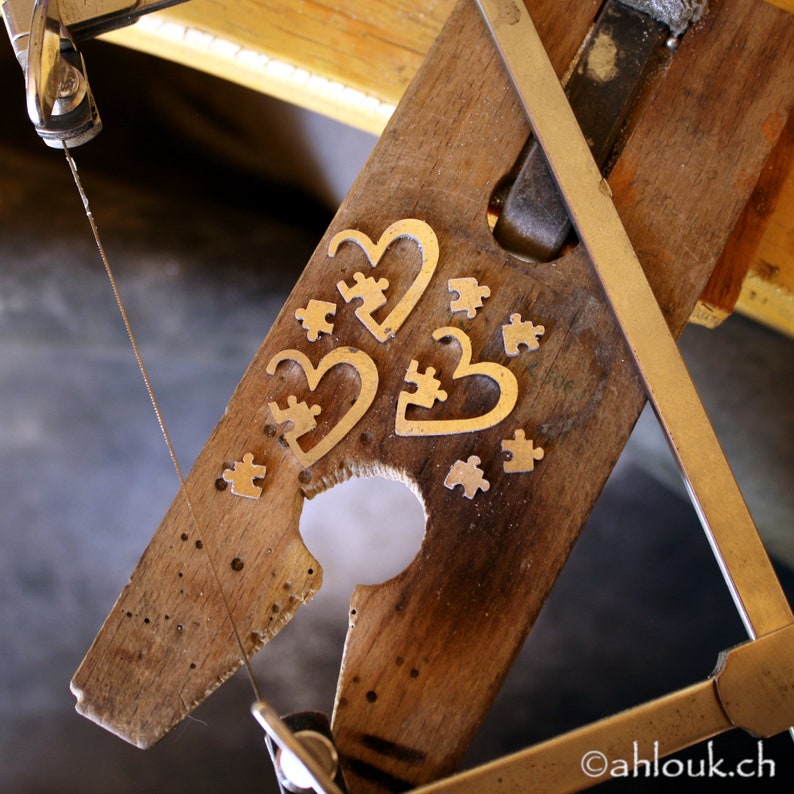 May include: A close-up of a wooden board with a jigsaw puzzle design cut out of it. The board is being cut with a saw. The puzzle pieces are in the shape of hearts and are made of metal.