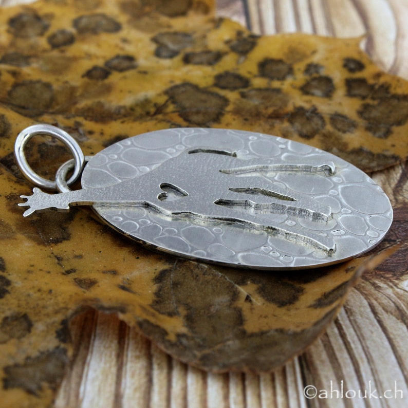May include: A silver pendant with an oval shape and a textured surface. The pendant features a cut-out design of a figure with outstretched arms and a heart shape. The pendant is attached to a silver ring.