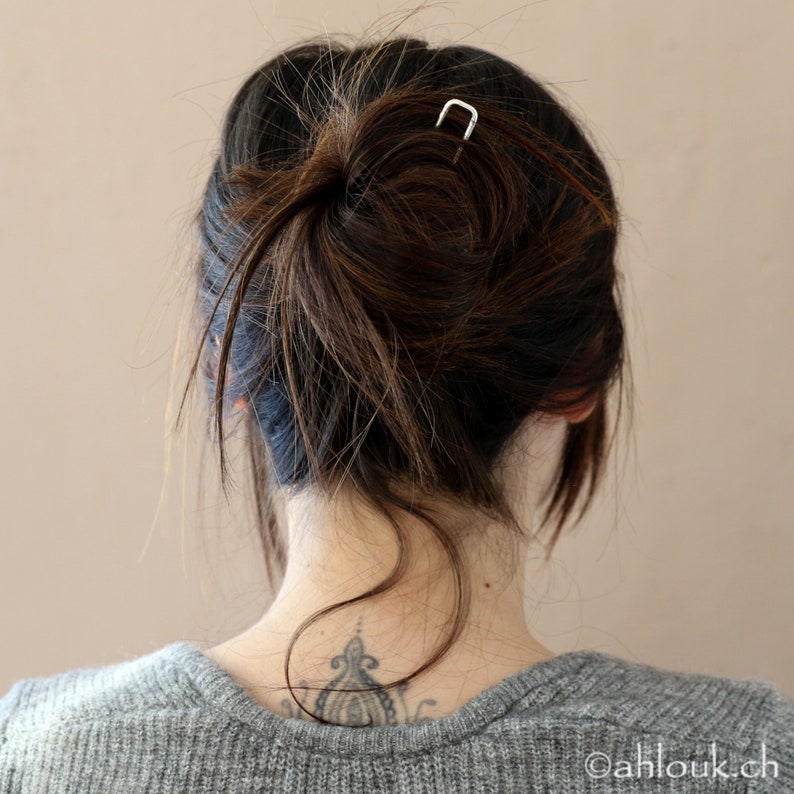 May include: A woman with long brown hair pulled back in a messy bun, secured with a silver hair clip. She is wearing a gray sweater and has a small tattoo on the back of her neck.