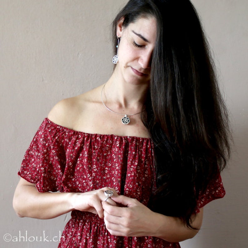May include: A woman wearing a red floral off-the-shoulder dress with a silver necklace and earrings featuring a circular design. She is wearing a silver ring on her finger.