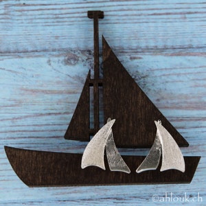 K&ouml;nnte beinhalten: Ein Paar silberne Segelschiff-Ohrringe mit strukturierter Oberfl&auml;che. Die Ohrringe werden auf einem ausgeschnittenen Holzsegelschiff pr&auml;sentiert.