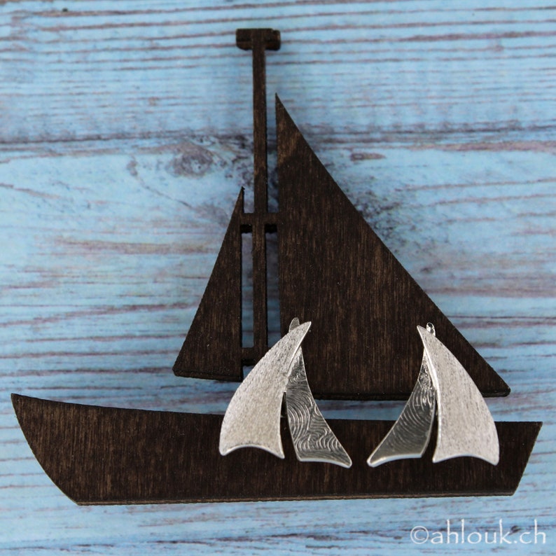 K&ouml;nnte beinhalten: Ein Paar silberne Segelschiff-Ohrringe mit strukturierter Oberfl&auml;che. Die Ohrringe werden auf einem ausgeschnittenen Holzsegelschiff pr&auml;sentiert.