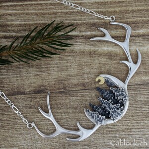 May include: A silver necklace with a crescent moon and a forest scene pendant. The pendant is made of textured silver and has a gold crescent moon. The necklace is on a silver chain.