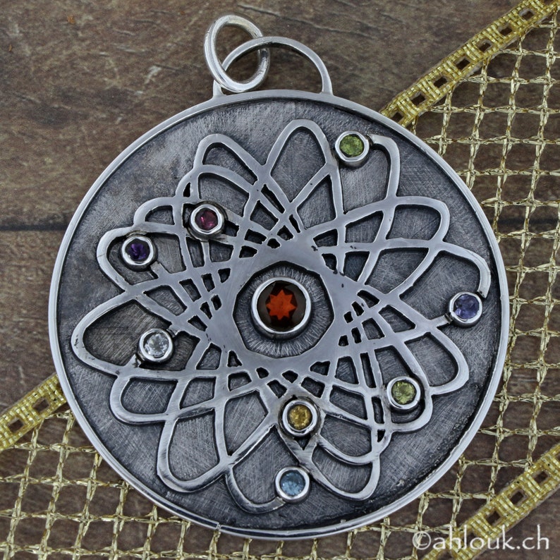 May include: A silver pendant with a circular design featuring a stylized atom with a central red gemstone surrounded by smaller gemstones in various colors.