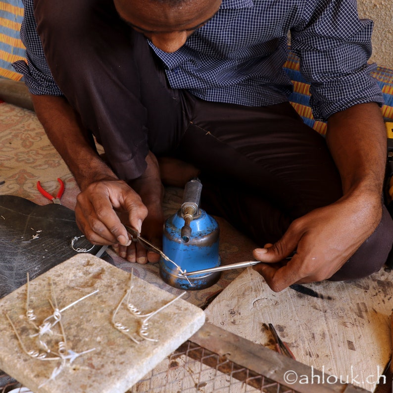 May include: A person is working on a piece of jewelry with a soldering iron and a small torch. The jewelry is on a wooden surface with a white tile. The person is wearing a blue and white plaid shirt.