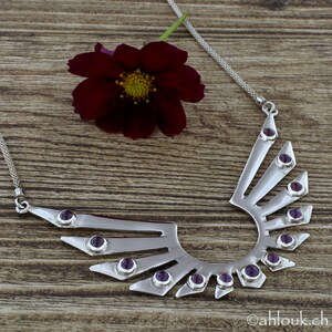 May include: A silver necklace with a geometric design featuring a crescent shape with radiating points. The necklace is adorned with purple gemstones.
