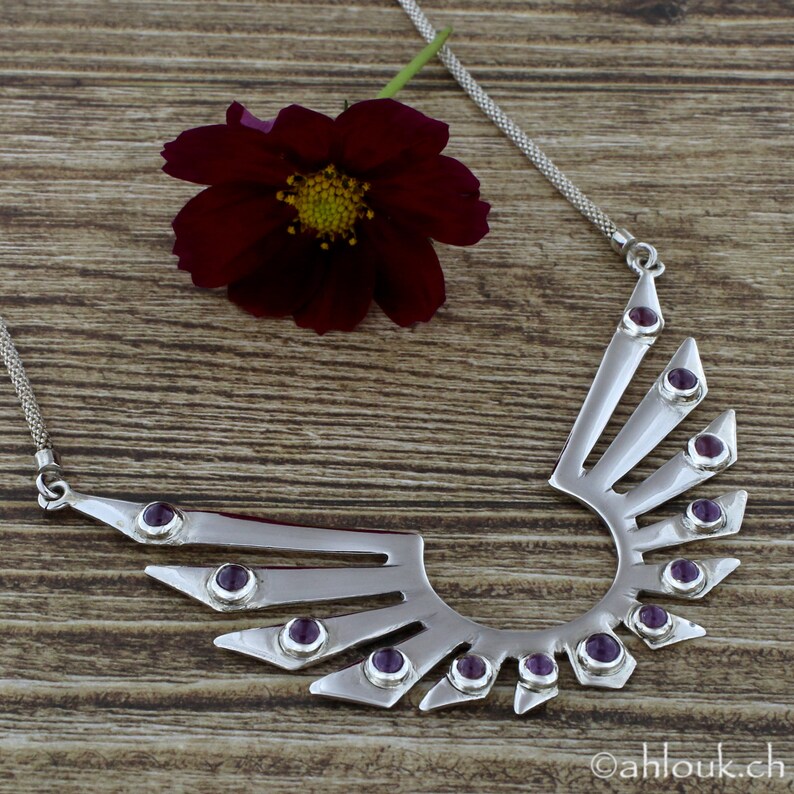 May include: A silver necklace with a geometric design featuring a crescent shape with radiating points. The necklace is adorned with purple gemstones.