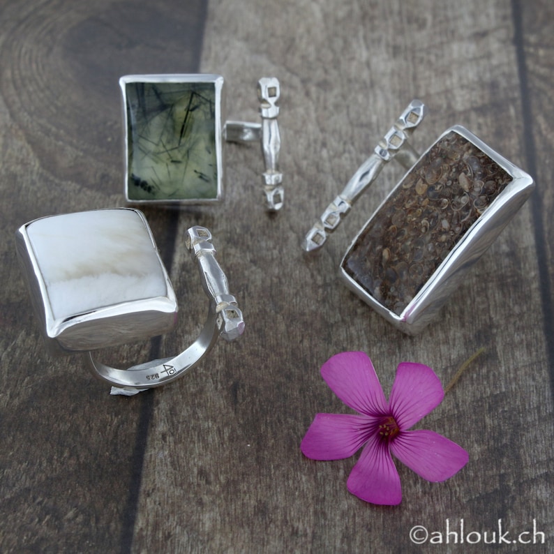 May include: Three silver rings with different stone inlays. The rings are on a wooden surface. The stones are a green and black stone, a brown stone, and a white stone.