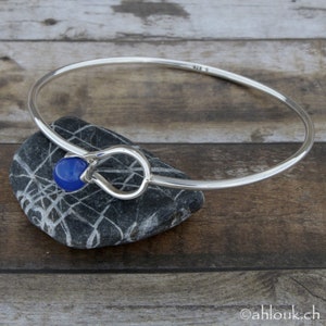 May include: A silver bangle bracelet with a blue gemstone accent. The bracelet is open and has a simple, elegant design.