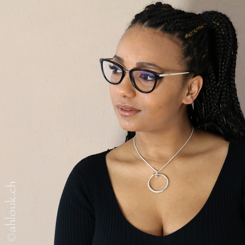 May include: A woman with dark brown braided hair wearing black cat-eye glasses and a black top with a silver necklace featuring a textured circle pendant.