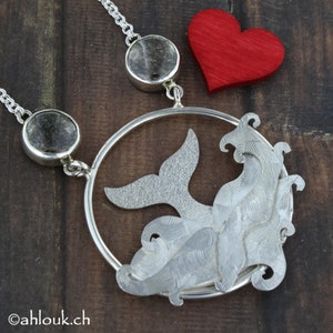 May include: A silver necklace with a whale tail pendant and two clear quartz stones. The pendant is a circle with a whale tail design and a textured surface. The necklace is on a wooden background.