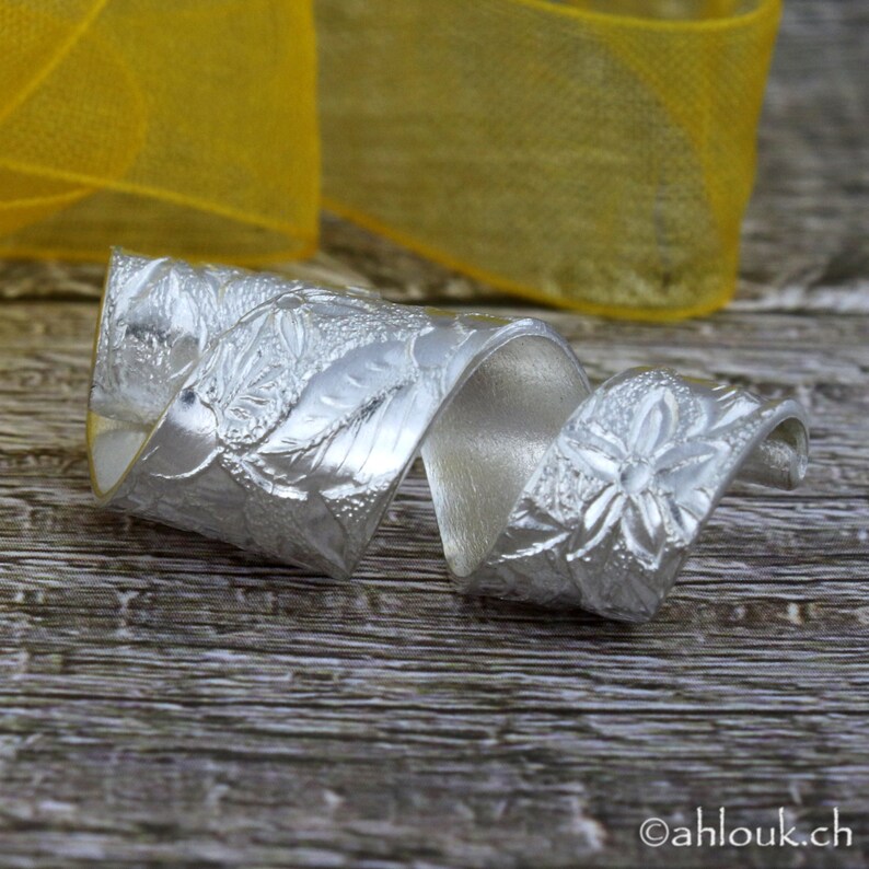 May include: A silver ring with a floral design, twisted into a spiral shape. The ring is on a wooden surface.
