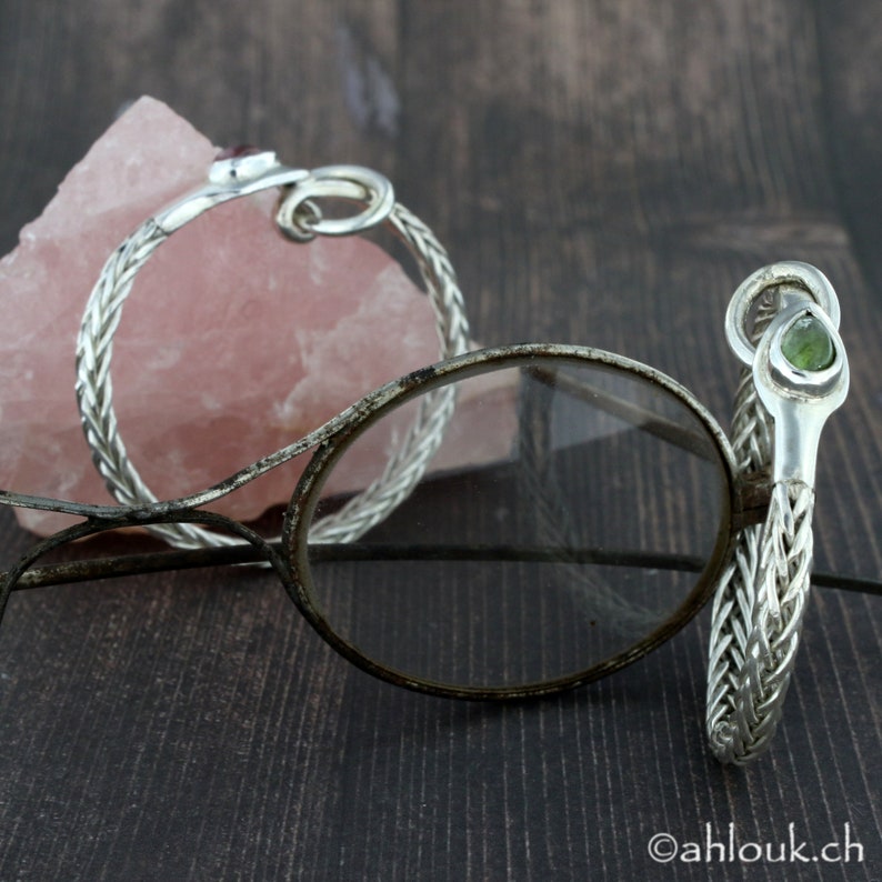 May include: Two silver bracelets with braided chain designs. One bracelet features a red gemstone, the other a green gemstone.