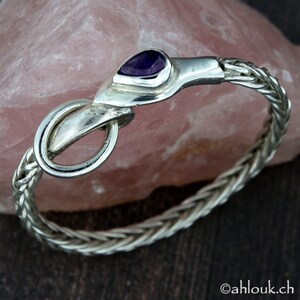 May include: A silver bracelet with a braided chain design and a purple gemstone clasp. The clasp is shaped like a snake's head.