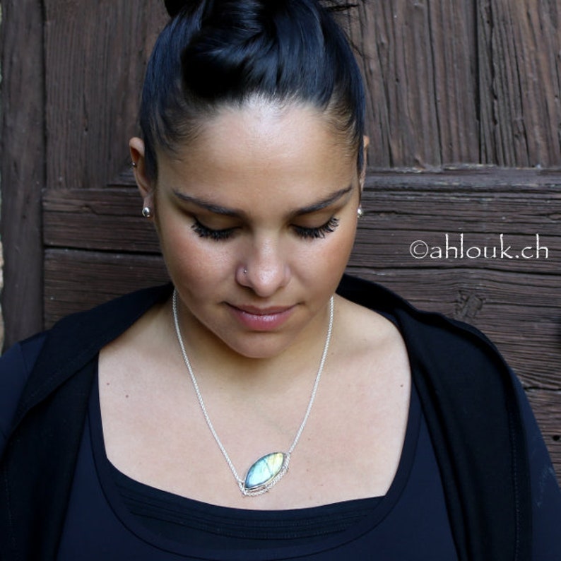 Puede incluir: Una mujer con una blusa negra y un collar de cadena plateada con un colgante de piedra preciosa ovalada iridiscente.