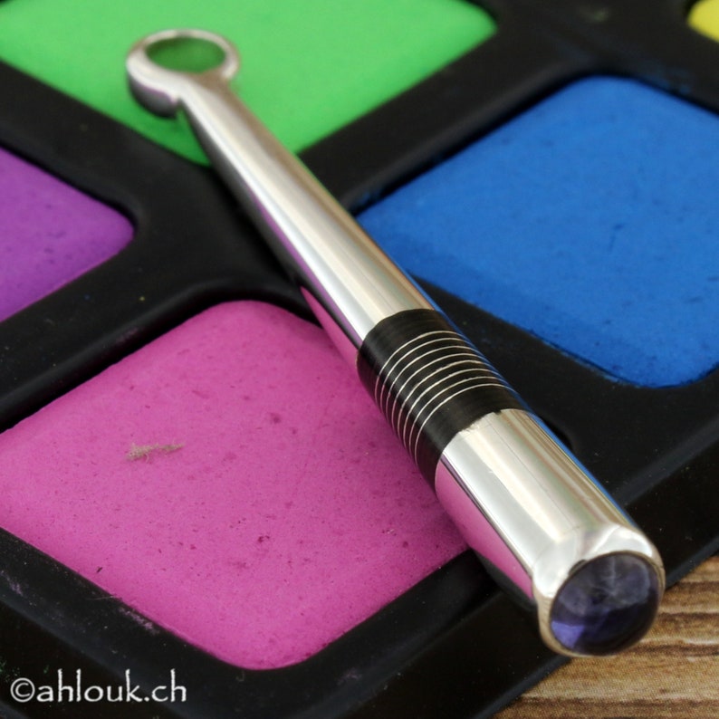 May include: A silver and black pen with a purple gemstone at the end. The pen is lying on a colorful palette of paint.