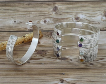 May include: Five silver bracelets with different colored gemstones. Each bracelet has a braided design and a single gemstone set into the metal. The gemstones are red, green, purple, blue, and orange.