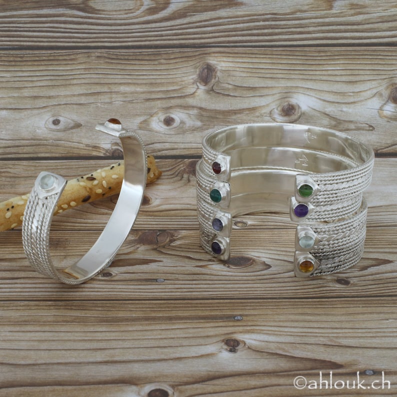 Puede incluir: Cinco pulseras de plata con piedras preciosas de diferentes colores. Cada pulsera tiene un dise&ntilde;o trenzado y una sola piedra preciosa engastada en el metal. Las piedras preciosas son roja, verde, morada, azul y naranja.
