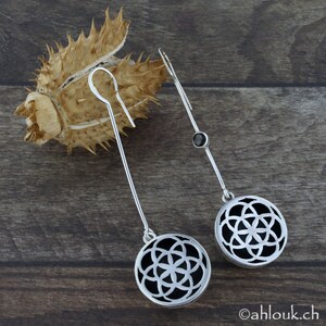 May include: A pair of silver dangle earrings with black geometric designs. Each earring features a round pendant with a cut-out pattern and a small black gemstone accent.
