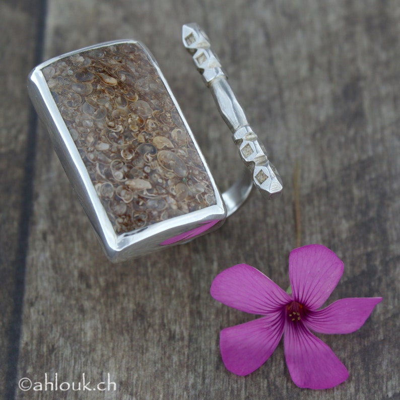 May include: A silver ring with a rectangular stone set in the centre. The stone is a light brown colour and has a pattern of small, round shapes. The ring is on a wooden surface with a pink flower.