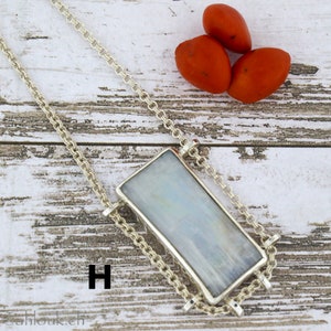Puede incluir: Un collar de cadena de plata con un colgante rectangular de piedra iridiscente. El colgante est&aacute; engastado en un marco de plata y cuelga de la cadena. El collar est&aacute; sobre una superficie de madera blanca con tres bayas rojas.