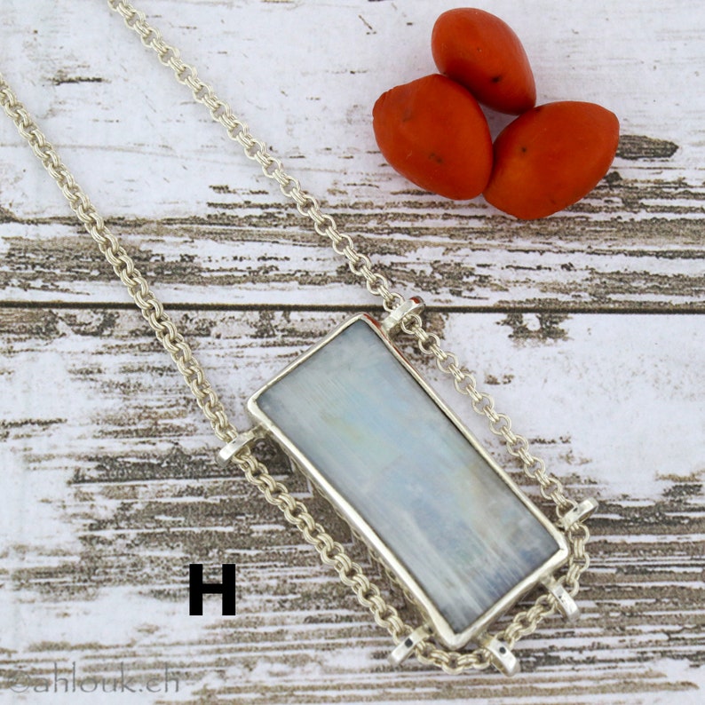Puede incluir: Un collar de cadena de plata con un colgante rectangular de piedra iridiscente. El colgante est&aacute; engastado en un marco de plata y cuelga de la cadena. El collar est&aacute; sobre una superficie de madera blanca con tres bayas rojas.