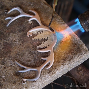 May include: A silver reindeer antler pendant with a cutout design of a forest is being soldered with a torch. The pendant is on a gray stone surface.