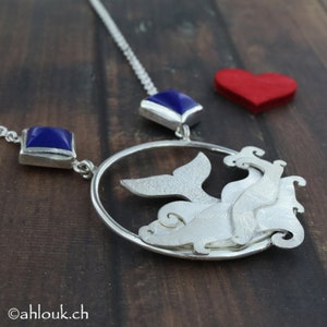 May include: Silver necklace with a whale tail pendant and two blue gemstone accents. The pendant is a circle with a whale tail inside and a textured design.