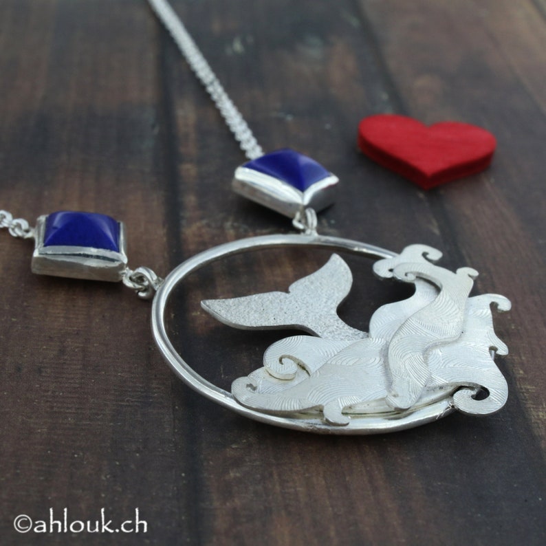 May include: Silver necklace with a whale tail pendant and two blue gemstone accents. The pendant is a circle with a whale tail inside and a textured design.