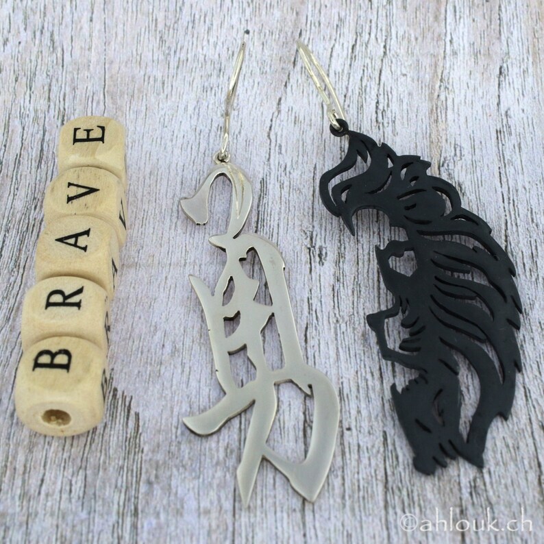 May include: A pair of silver earrings with a cutout design. One earring features the word "brave" in Japanese characters, while the other earring is a stylized black silhouette of a wolf's head.