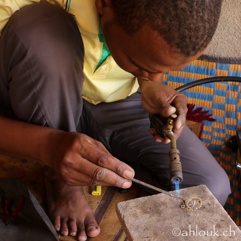 May include: A person uses a blowtorch to heat a piece of metal jewelry. The metal is glowing orange hot. The person is wearing a yellow shirt and dark pants. The background is a colorful patterned rug.