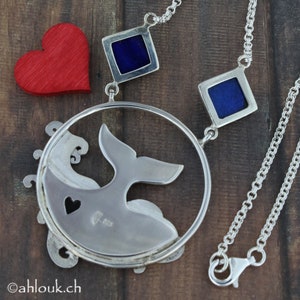 May include: A silver necklace with a whale tail pendant and two blue enamel squares. The pendant is attached to a silver chain with a lobster clasp.