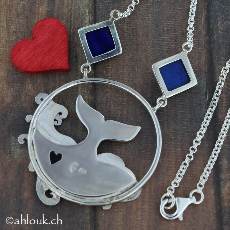 May include: A silver necklace with a whale tail pendant and two blue enamel squares. The pendant is attached to a silver chain with a lobster clasp.