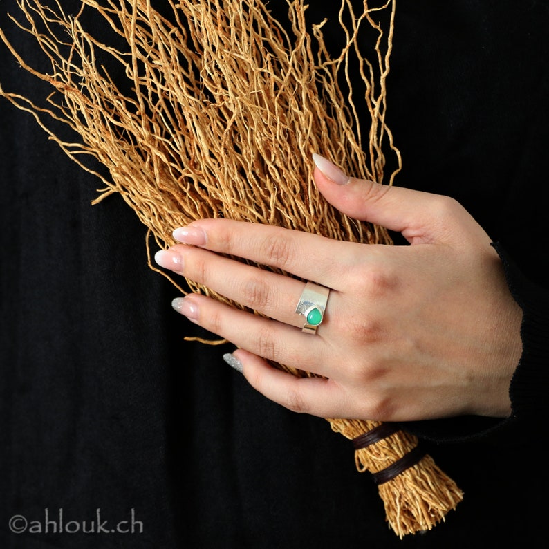 May include: A silver ring with a green gemstone set in a textured band. The ring is worn on a hand holding a bundle of dried twigs.