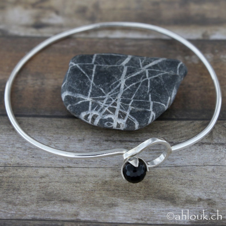May include: A silver bangle bracelet with a black onyx stone accent. The bracelet is open and has a simple, elegant design.