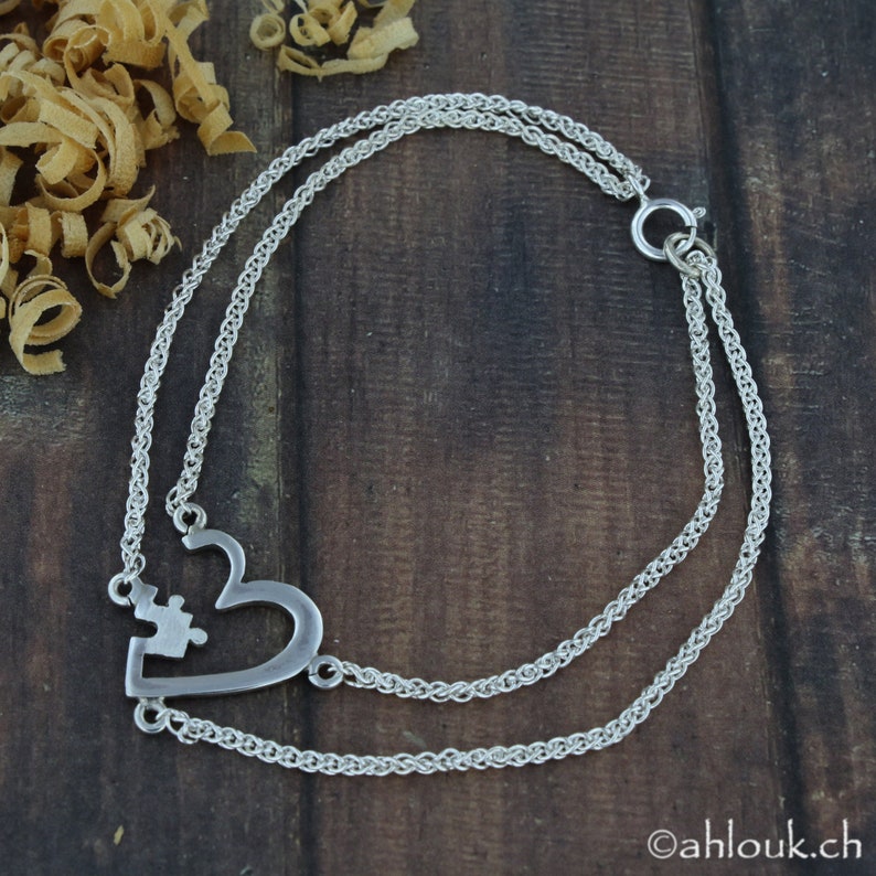 May include: A silver chain bracelet with a heart-shaped charm featuring a puzzle piece cutout. The chain is double-stranded and has a lobster clasp.