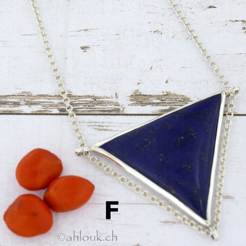 Puede incluir: Un collar de cadena de plata con un colgante triangular que presenta una piedra de lapisl&aacute;zuli azul oscuro. El colgante est&aacute; engastado en un marco de plata y tiene un dise&ntilde;o de cadena doble. El collar est&aacute; fotografiado sobre una superficie de madera clara con tres peque&ntilde;as semillas naranjas.