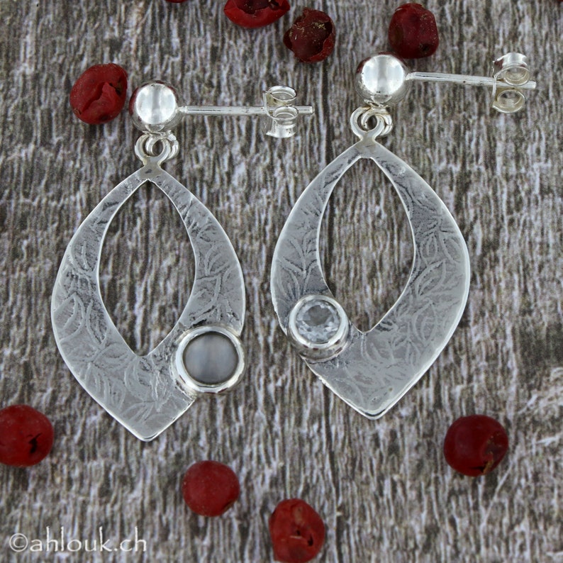 May include: A pair of silver earrings with a teardrop-shaped design. Each earring features a small, clear stone set in the center of the teardrop. The earrings have a textured surface and are accented with a small, round silver bead at the top.
