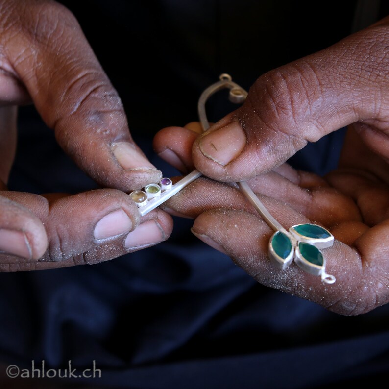 May include: A silver necklace with green gemstone leaves and a row of colorful gemstones. The necklace is being held by a person's hands.