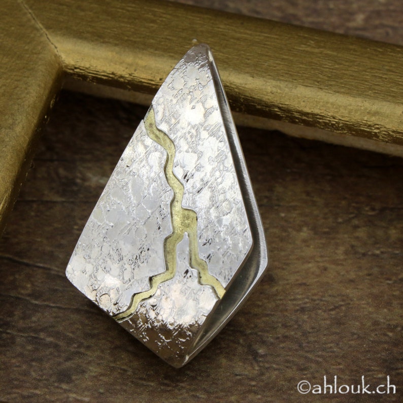 May include: A silver-toned pendant with a textured surface and a gold-colored, meandering inlay. The pendant has a geometric, diamond-like shape. The background is a blurred wooden surface.