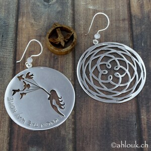 May include: A pair of silver earrings. One earring features a circular design with a cutout pattern. The other earring features a cutout design of a girl holding a string of birds with the text "I know the way home".