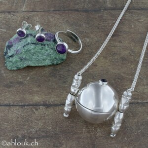 May include: A set of silver jewelry featuring amethyst gemstones. The set includes a pair of earrings, a ring, and a pendant necklace with a round, hinged locket. The locket is decorated with a silver chain and a silver ball.